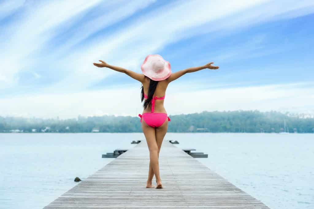 Imagem de uma mulher usando um biquíni rosa e um chapéu na cor rosa. Ela está caminhando sobre um píer próximo a um grande lago. Ela está tendo uma sensação de liberdade, pois está com os braços abertos, respirando um ar puro.