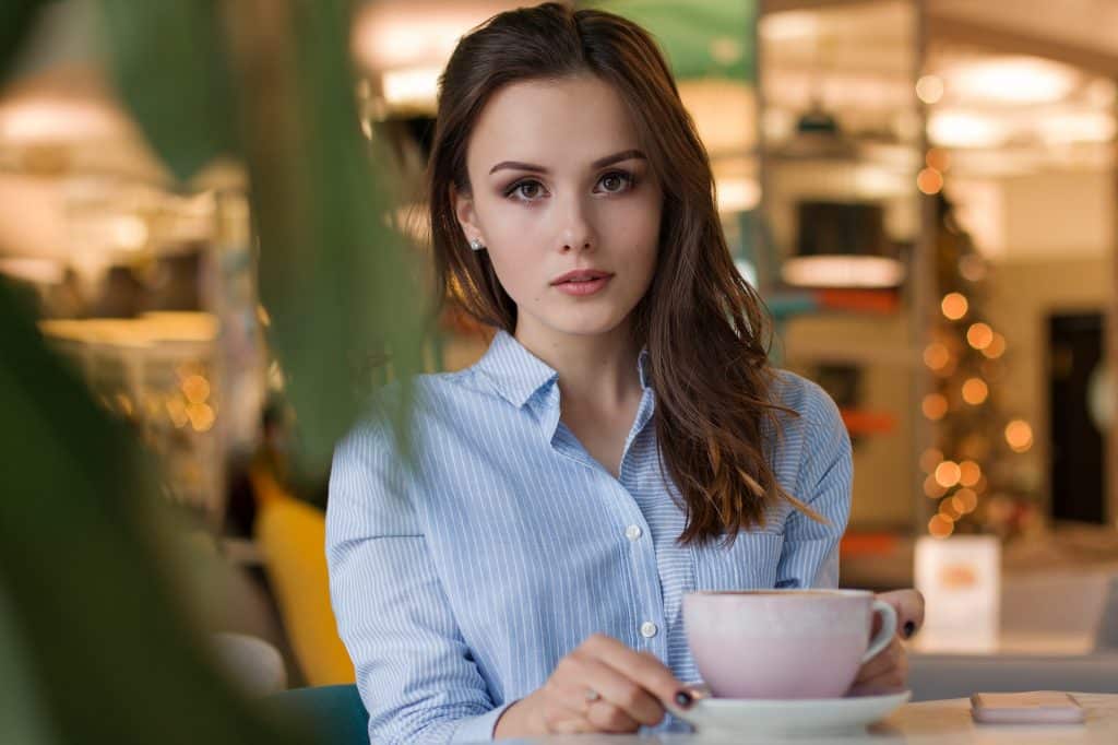 Imagem de uma linda mulher sentada em uma mesa de um café. Ela usa uma camisa azul clara com listras brancas e segura uma linda e grande caneca de porcelana.