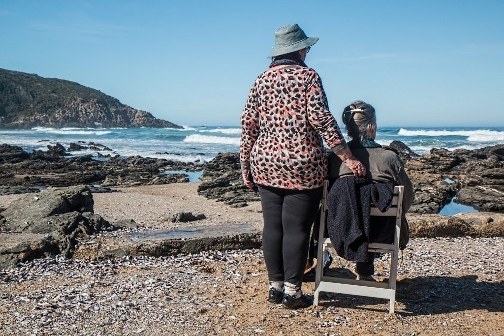 Imagem de duas amigas em uma praia. Uma está sentada em uma cadeira de madeira e a outra está em pé, apoiando a outra. Ambas olham para as águas e ondas do mar.