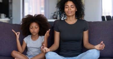 Mulher adulta e menina sentadas em sofá. Elas praticam meditação.