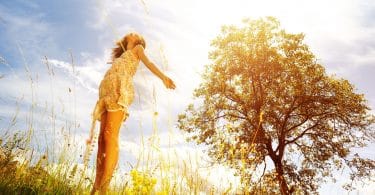 Mulher com os braços abertos e cabeça erguida. O cenário é externo, de vegetação e solar.