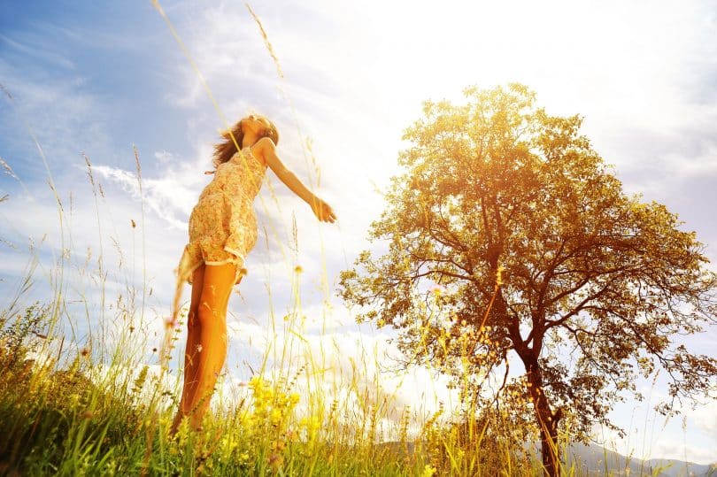 Mulher com os braços abertos e cabeça erguida. O cenário é externo, de vegetação e solar.