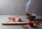 Imagem de fundo cinza e em destaque temos um incensário com um incenso de massala aceso, algumas flores espalhadas sobre a mesa e um pote de ferro.