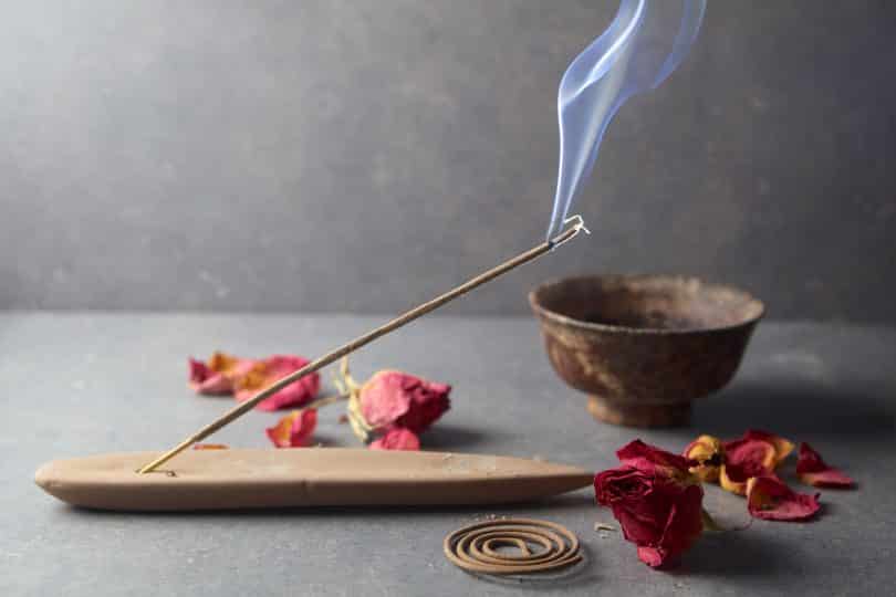 Imagem de fundo cinza e em destaque temos um incensário com um incenso de massala aceso, algumas flores espalhadas sobre a mesa e um pote de ferro.