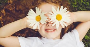 Menina branca segurando flores em seus olhos enquanto sorri.