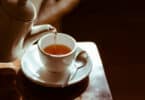 Bule de chá despejando seu conteúdo em uma xícara que repousa sobre a ponta de uma mesa.