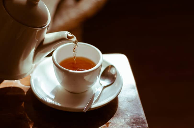 Bule de chá despejando seu conteúdo em uma xícara que repousa sobre a ponta de uma mesa.