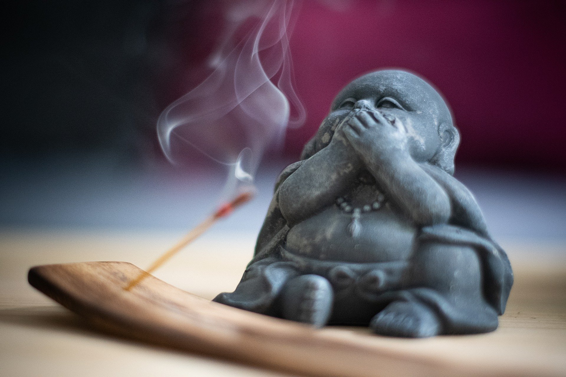 Imagem de um incensário com um incenso de massala aceso. Ao lado do incensário uma pequena estátua do Buda com as duas mãos sobre a boca.
