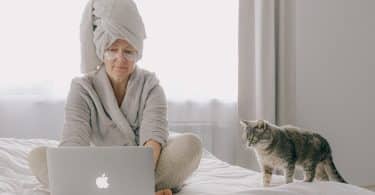 Senhora sentada na cama usando seu laptop ao lado de seu gatinho