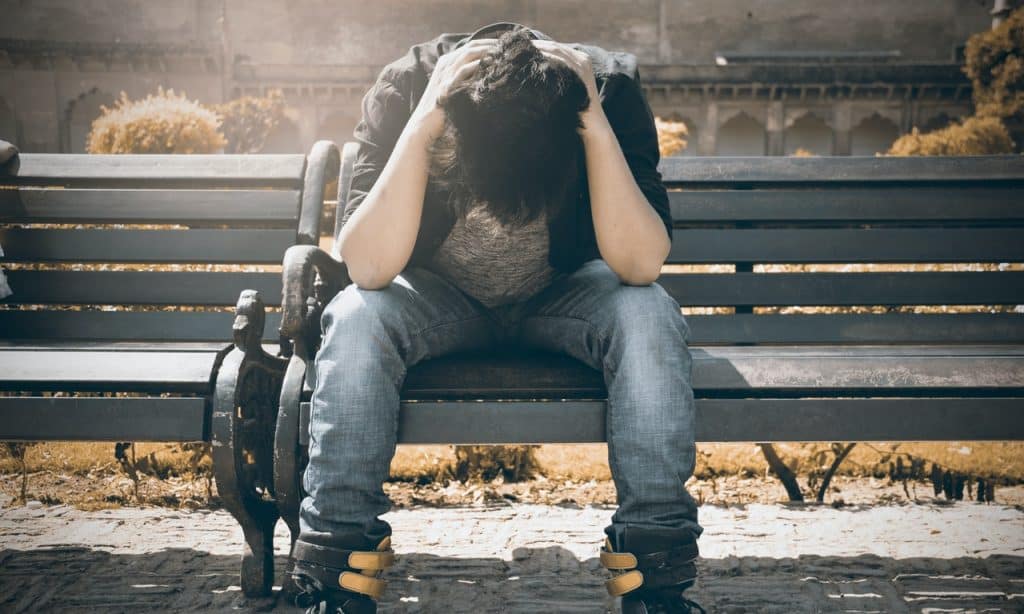 Homem sentado em banco de praça. Ele apoia os cotovelos nos joelhos e tem as mãos sobre a nuca.
