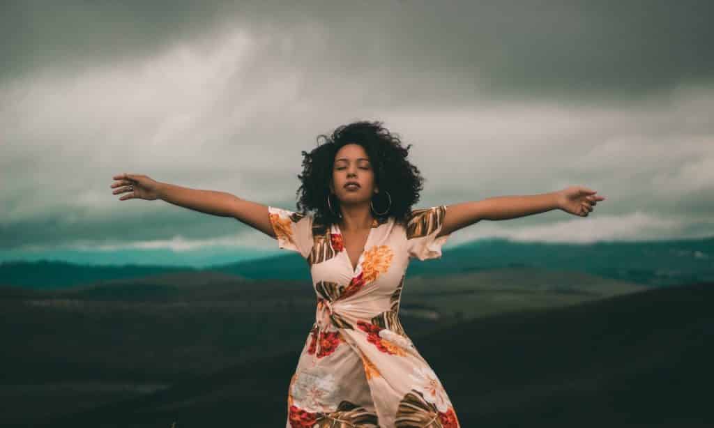 Mulher com os braços abertos e erguidos em ambiente externo e montanhoso. 