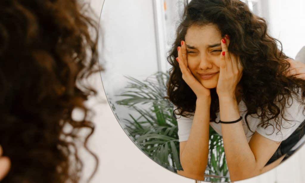 Mulher observa seu próprio reflexo no espelho. Seu semblante é aflito.