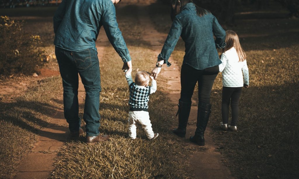 Família composta por duas crianças e dois adultos caminha em ambiente externo. 