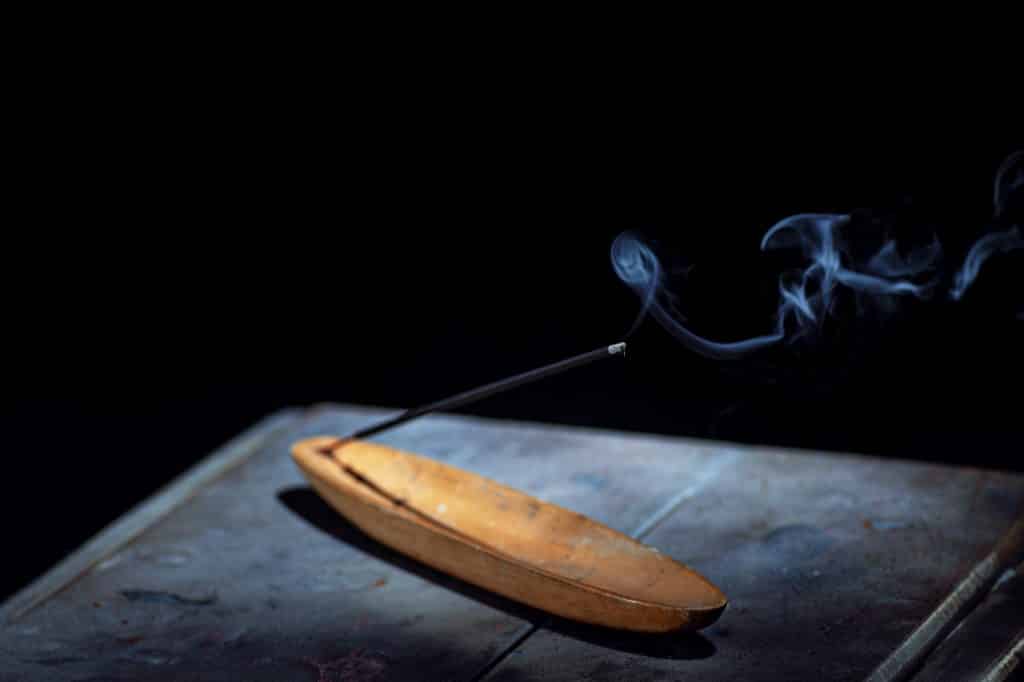 Imagem de fundo preto e em destaque um incensário de madeira e dentro dele um incenso de sálvia sendo queimado. O incensário está sobre uma mesa de madeira.
