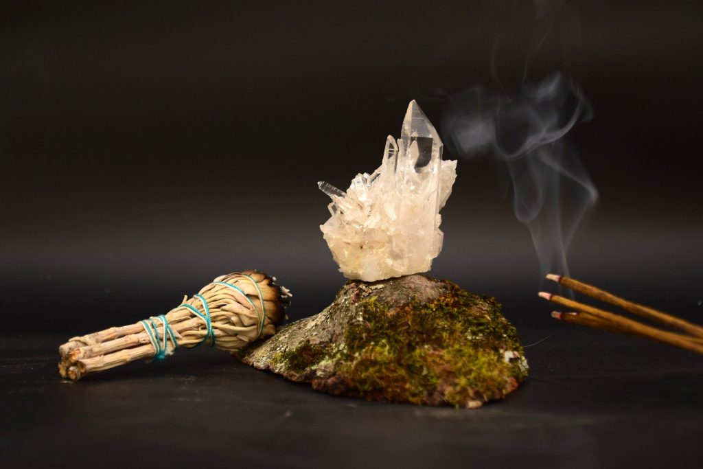 Imagem de um pedra e sobre ela um lindo cristal branco. Do lado direito da pedra temos quatro incensos palitos de sálvia sendo queimados e do lado esquerdo, vários incensos já queimados.
