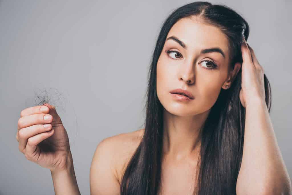 Imagem de uma mulher de cabelos longos. Uma das suas mãos está sobre a cabeça e a outra segurando um monte de fios de cabelo que acabaram de cair.