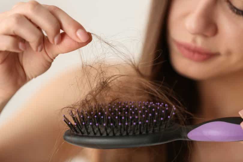 Imagem de um mulher segurando uma escova de cabelo na cor preto com cabo roxo. Com uma das mãos ela tira da escova vários fios de cabelo.