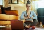 Mulher com roupas de academia sentada no chão de uma casa meditando usando o laptop