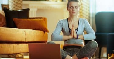 Mulher com roupas de academia sentada no chão de uma casa meditando usando o laptop