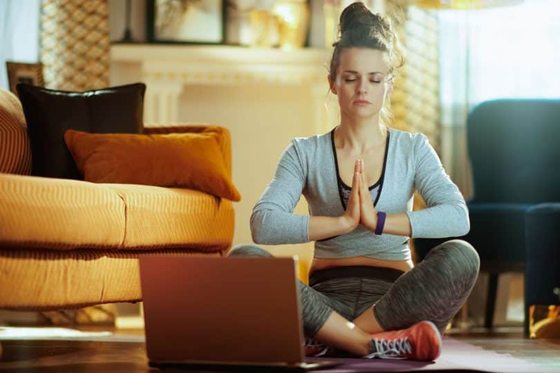 Mulher com roupas de academia sentada no chão de uma casa meditando usando o laptop