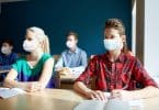 Grupo de estudantes com máscaras. Eles estão em uma sala de aula, sentados à mesa, que contém cadernos e livros.