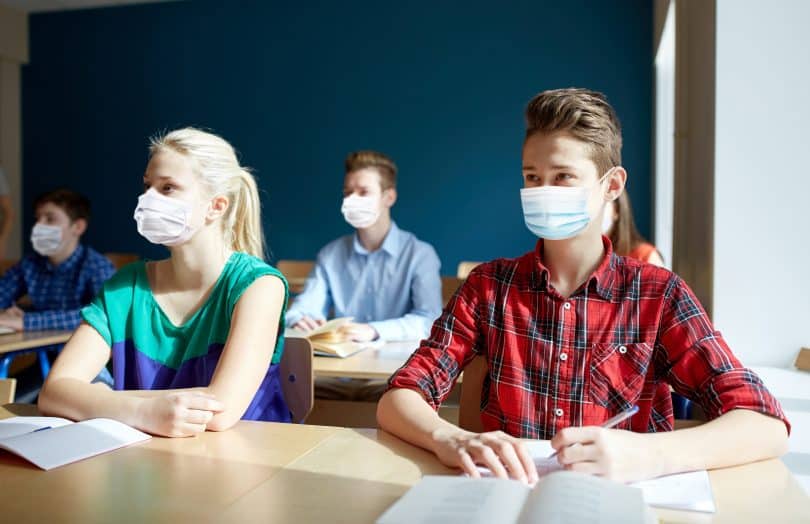 Grupo de estudantes com máscaras. Eles estão em uma sala de aula, sentados à mesa, que contém cadernos e livros.