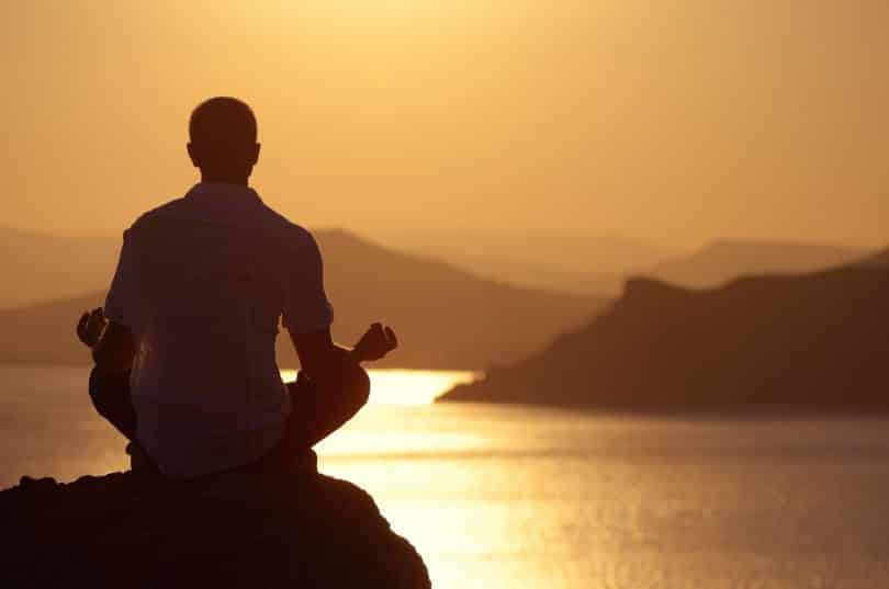 Homem medita sobre pedra. Ao fundo, há o mar, montanhas e o cenário é de pôr do sol.