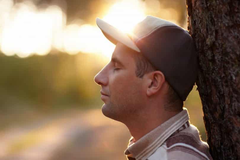 Foto de perfil de um homem com os olhos fechados e encostado em uma árvore.