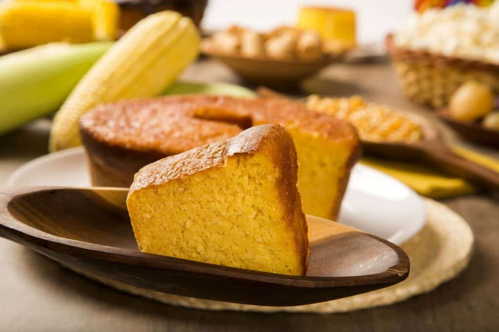 Fatia de bolo de milho cortada à frente do bolo em si. Ao fundo, há espigas de milhos e outros alimentos.