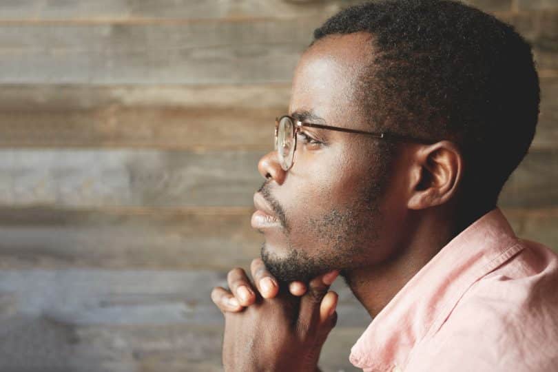 Homem jovem visto de perfil observa o horizonte. Ele apoia o queixo sobre as duas mãos unidas. O fundo é de madeira.