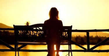Mulher sentada em banco assiste ao pôr do sol.