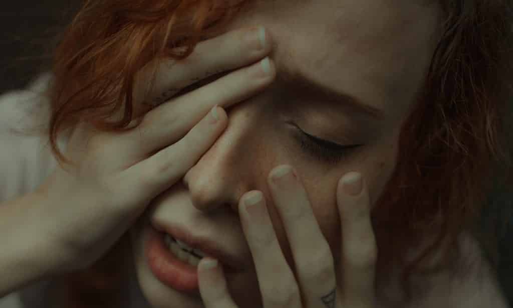 Mulher com as mãos sobre o rosto e olhos fechados. Seu semblante é aflito.