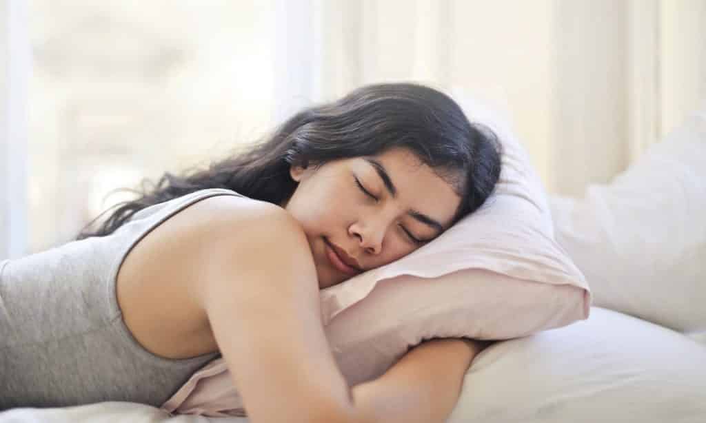 Mulher de bruços deitada em cama dorme. Seus braços estão sob o travesseiro.