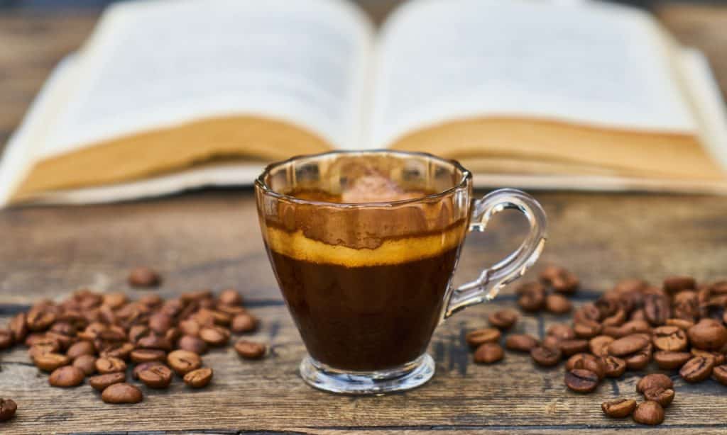 Xícara de café sobre superfície de madeira, onde também há grãos de café e um livro aberto ao fundo.