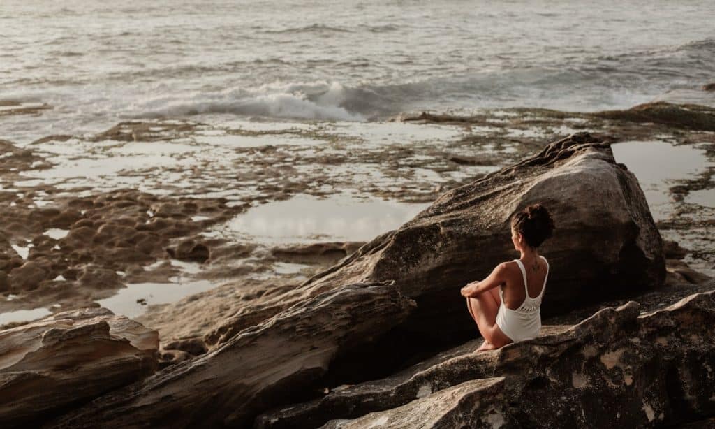 Mulher sentada em pedras à frente do mar.