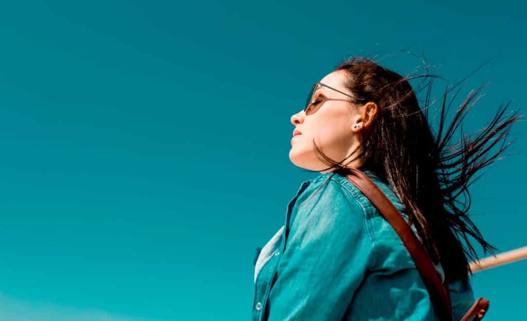 Mulher de olhos fechados e óculos escuros em ambiente externo. Ela é vista de perfil e seus cabelos voam ao vento.