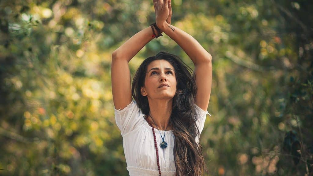 Mulher olha para cima, onde suas mãos se unem, erguidas. Ela está em cenário externo.