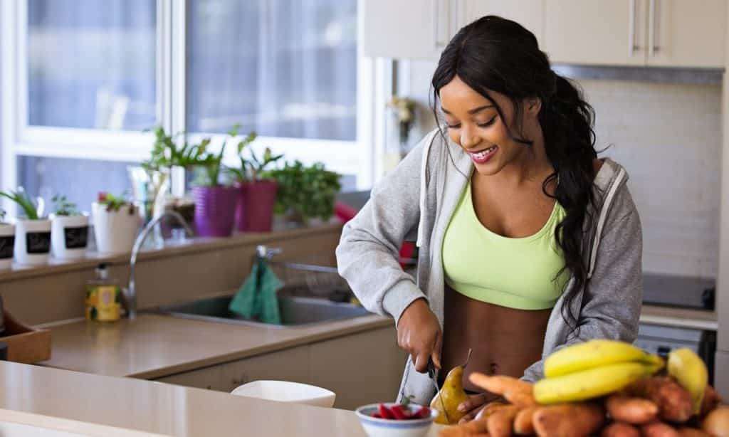 Mulher corta fruta sobre bancada. Ela está em uma cozinha.