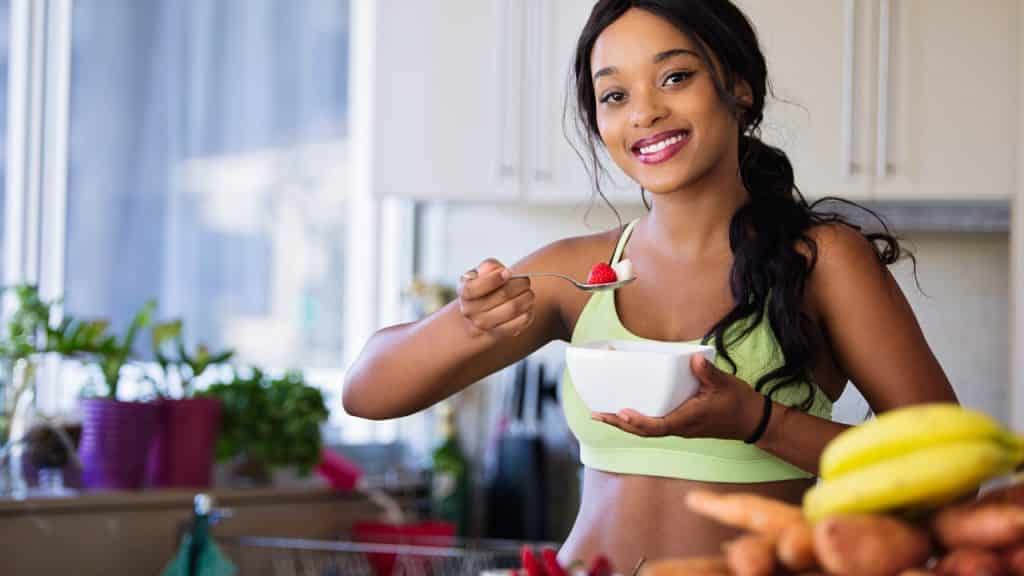 Mulher segura uma tigela de porcelana branca e uma colher com frutas. Ela sorri e intenciona comer as frutas. O cenário é de uma cozinha.