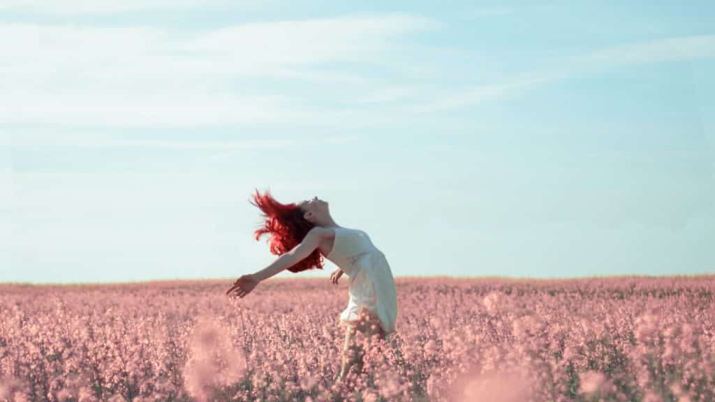 Mulher, com os braços abertos, inclina-se para trás em campo de flores. Seus cabelos estão ao vento.