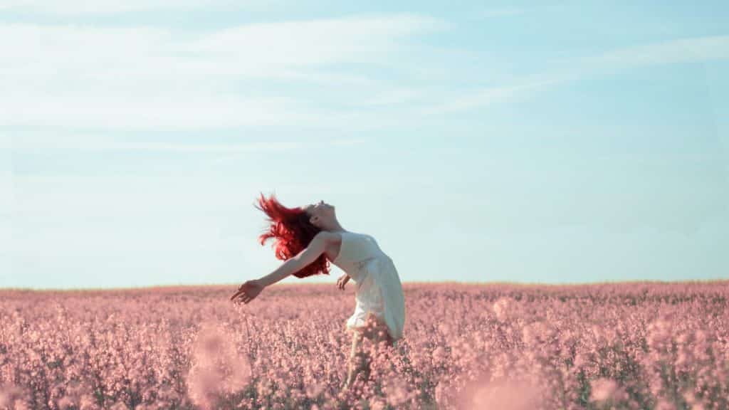 Mulher, com os braços abertos, inclina-se para trás em campo de flores. Seus cabelos estão ao vento. 