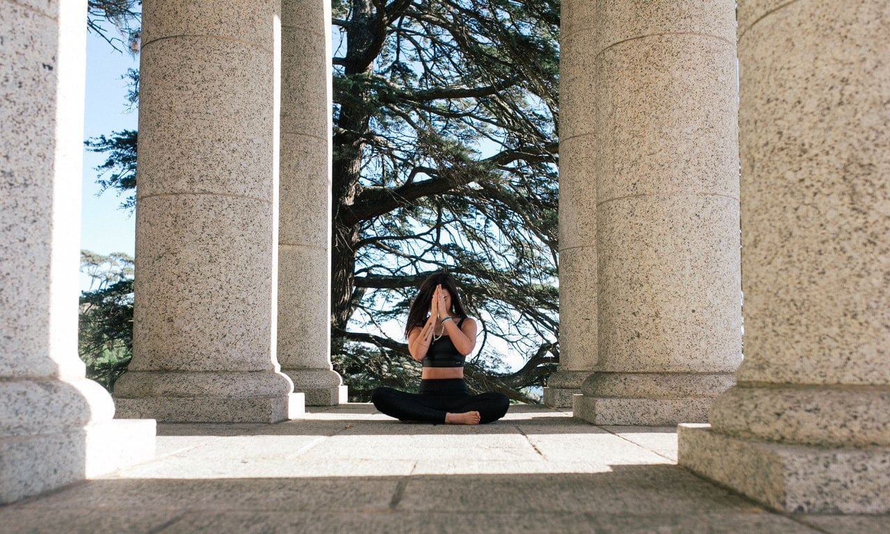 Mulher medita em área externa de colunas de concreto cinza.
