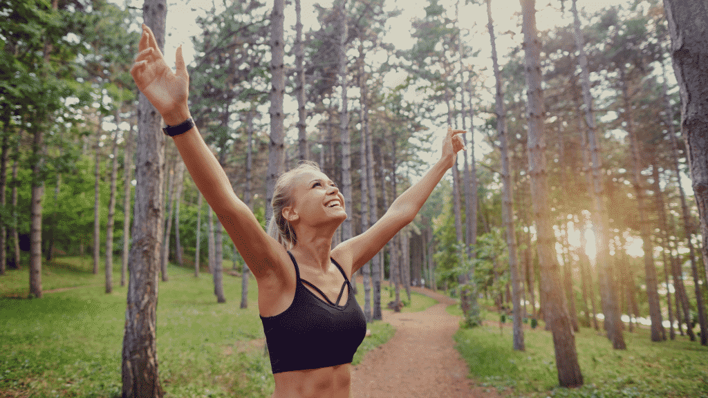 Mulher sorrido com os braços erguidos na floresta depois de correr