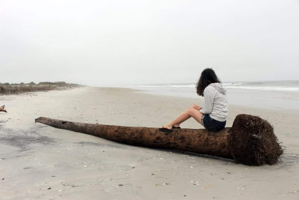 Mulher branca sentada no tronco de uma árvore na praia.