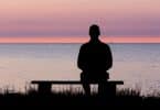 Homem sentado em banco à beira de lago observa o pôr do sol.