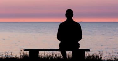 Homem sentado em banco à beira de lago observa o pôr do sol.