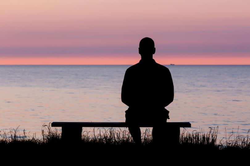 Homem sentado em banco à beira de lago observa o pôr do sol.