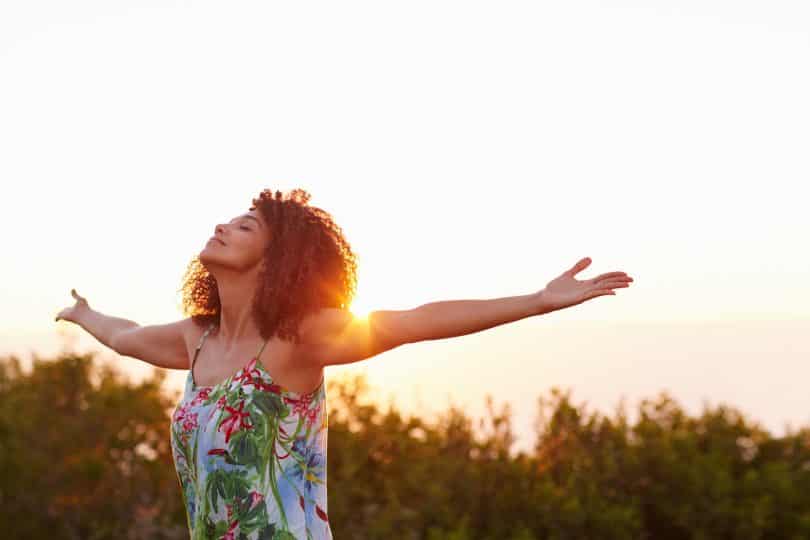 Mulher com os braços abertos e erguidos em ambiente externo. Sua cabeça está inclinada para trás e os olhos estão fechados.