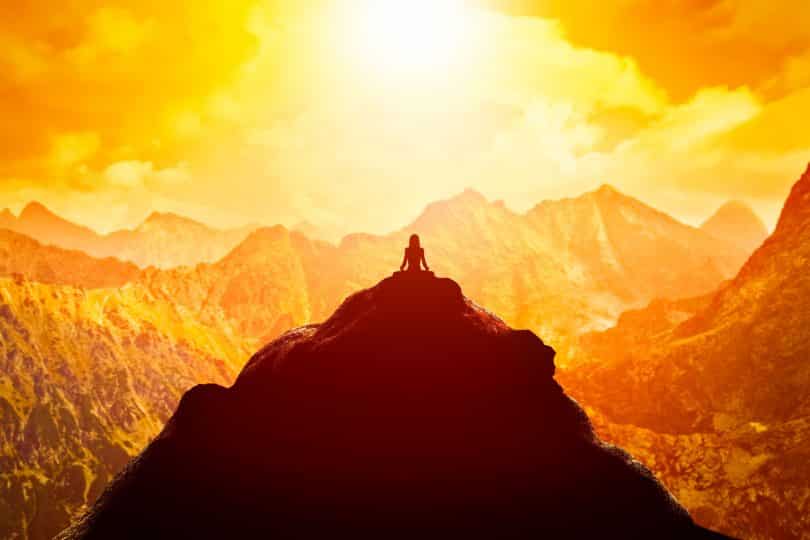 Mulher meditando em posição sentada de ioga no topo de uma montanha acima das nuvens ao pôr do sol.