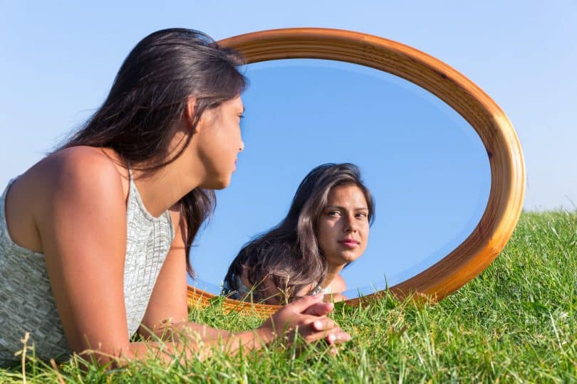 Mulher deitada de bruços na grama observa seu reflexo em espelho.